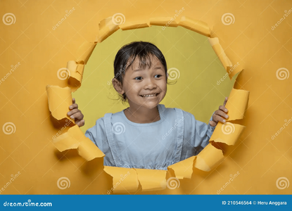 Cute little asian girl happy smiling poses torn yellow paper hole looking something aside chee